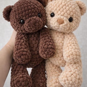 May include: Two plush teddy bears made of chunky, textured yarn. One bear is dark brown with black eyes, and the other is light beige with black eyes and a small white pom-pom detail on its head. Both bears have rounded ears and limbs.