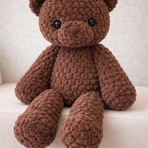 May include: A brown teddy bear crafted from chunky, textured yarn. The stuffed animal features a rounded head with small ears, a prominent snout, and two smooth, dark, circular eyes. Its body has long arms and legs with a consistent, nubby stitch pattern.