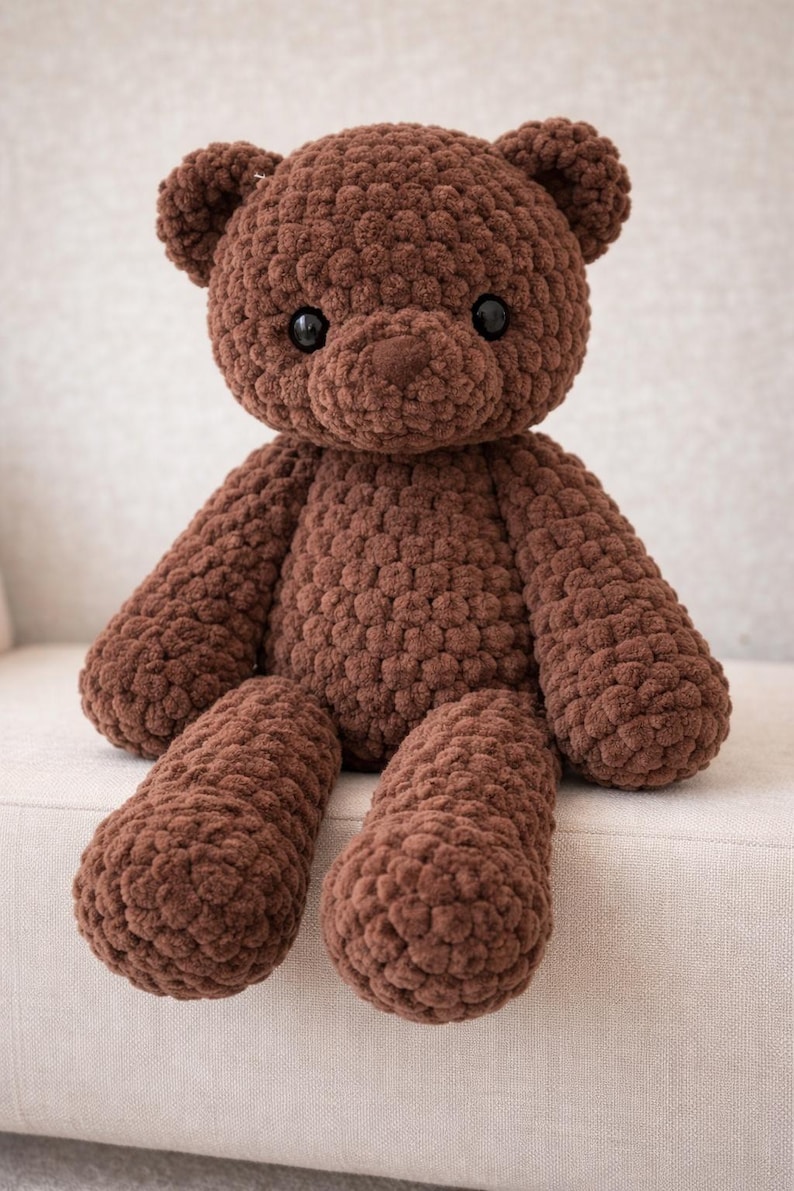 May include: A brown teddy bear crafted from chunky, textured yarn. The stuffed animal features a rounded head with small ears, a prominent snout, and two smooth, dark, circular eyes. Its body has long arms and legs with a consistent, nubby stitch pattern.