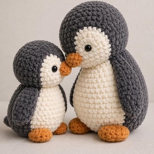Puede incluir: Dos peluches de pingüino hechos a mano en ganchillo de diferentes tamaños, ambos con cuerpos de color gris oscuro, vientres de color crema, pequeños picos naranjas y pies naranjas. El pingüino más grande está detrás del más pequeño.