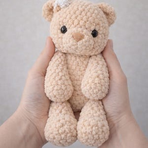 May include: A small, beige, crocheted teddy bear made from soft, textured yarn. The bear features two round black eyes, a small brown stitched nose, and a tiny white crocheted flower accessory attached to the top of its right ear.