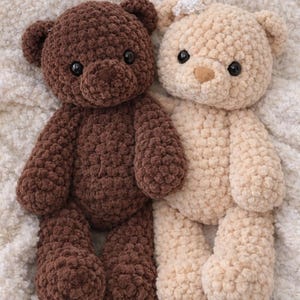 May include: Two plush teddy bears made of chunky, textured yarn. One bear is dark brown, and the other is cream-colored with a small white crocheted flower detail on its head. Both bears feature round black eyes and stitched noses.