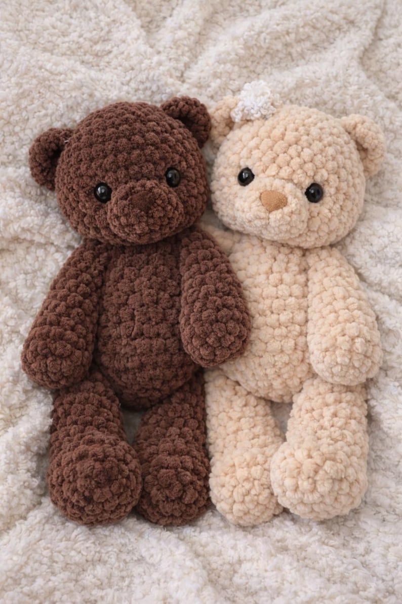 May include: Two plush teddy bears made of chunky, textured yarn. One bear is dark brown, and the other is cream-colored with a small white crocheted flower detail on its head. Both bears feature round black eyes and stitched noses.