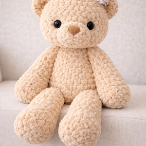 May include: A beige crochet teddy bear with a textured, bumpy surface, featuring two round black eyes, a brown stitched nose, and a small white crocheted flower accessory attached to the top of its head near the right ear.
