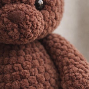 May include: A brown teddy bear crafted from thick, textured yarn. The plush toy features a rounded head, a prominent snout, and a single visible dark, glossy plastic eye. The surface of the bear has a distinct, bumpy, crocheted appearance.