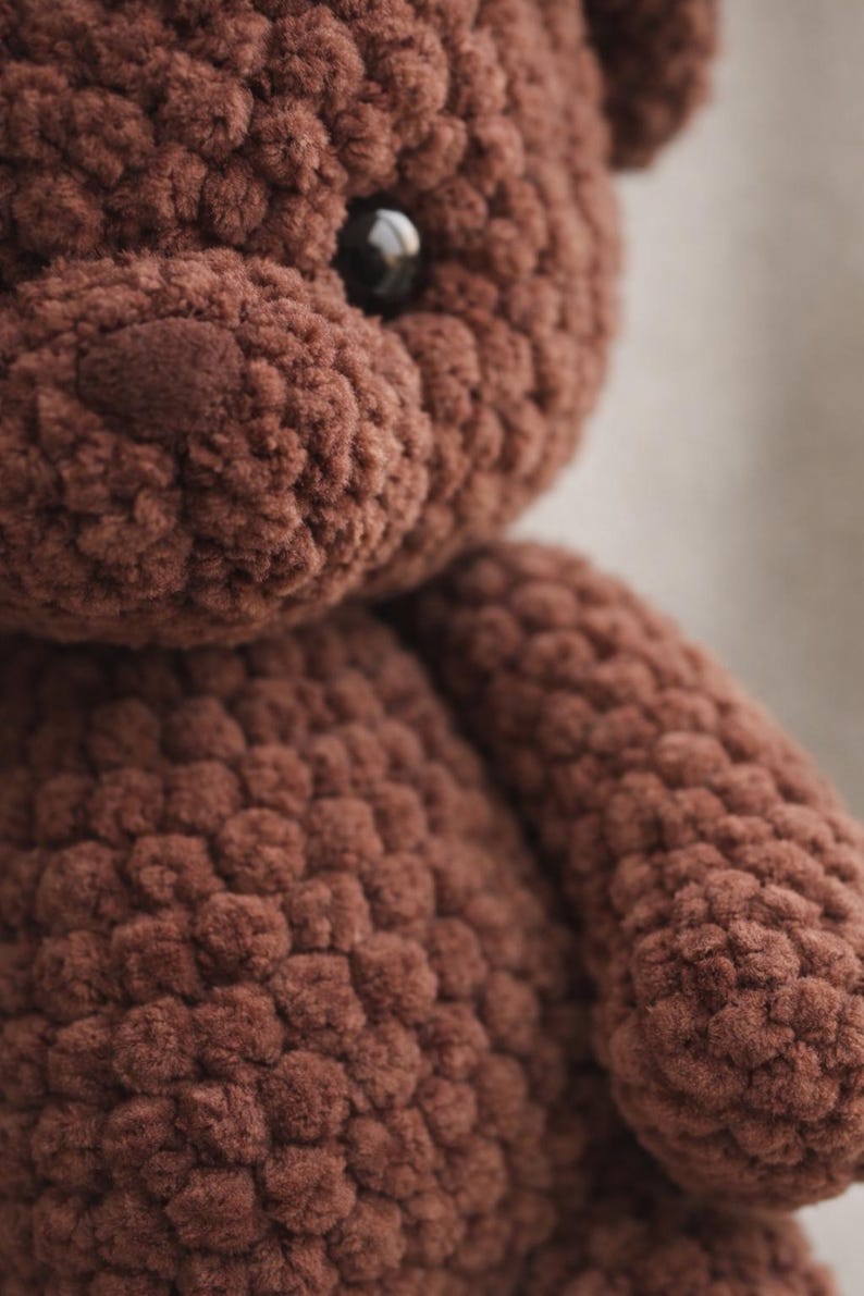 May include: A brown teddy bear crafted from thick, textured yarn. The plush toy features a rounded head, a prominent snout, and a single visible dark, glossy plastic eye. The surface of the bear has a distinct, bumpy, crocheted appearance.