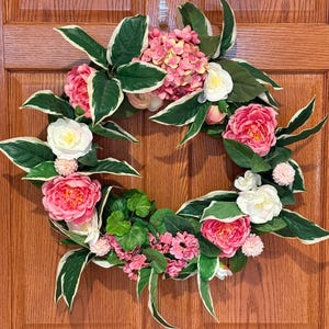 May include: A vibrant floral wreath featuring pink peonies, white roses, and pink hydrangeas, accented by green and white variegated leaves. This decorative wreath is perfect for adding a touch of spring to any door or wall.