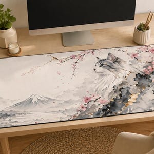 May include: A rectangular desk mat featuring a watercolor illustration of a cat with a mountain landscape and cherry blossoms. The cat is gray and white, with pink flowers and a mountain in the background. The mat is on a wooden desk.