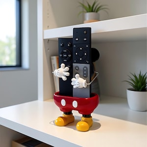 Puede incluir: Un soporte para mando a distancia novedoso con forma de personaje de dibujos animados. El soporte es rojo con zapatos amarillos y manos enguantadas blancas. Dos mandos a distancia negros se sujetan.