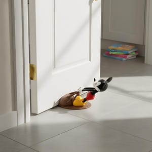 Könnte beinhalten: Ein skurriler Mickey Mouse Türstopper in Schwarz, Rot, Gelb und Braun. Der Türstopper ist so gestaltet, dass es aussieht, als würde Mickey die Tür offen halten. Der Türstopper steht auf einem hellgrauen Fliesenboden.