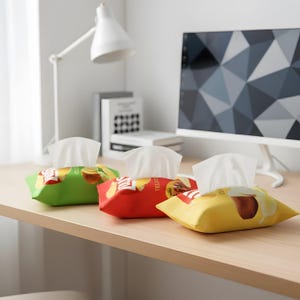 Puede incluir: Tres cajas de pañuelos de colores, verde, rojo y amarillo, están sobre un escritorio de madera. Cada caja tiene un pañuelo blanco que sobresale de la parte superior. Las cajas tienen gráficos y texto con temática de alimentos. Una lámpara de escritorio blanca y un monitor de ordenador están en el fondo.