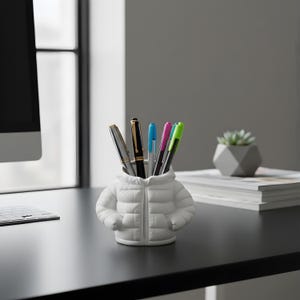 Puede incluir: Un portalápices de cerámica blanca con forma de chaqueta acolchada, con bolígrafos y rotuladores en su interior. El portalápices tiene una cremallera y dos bolsillos. Los bolígrafos son negros, plateados, dorados, azules, rosas y verdes. El portalápices está sobre un escritorio negro.