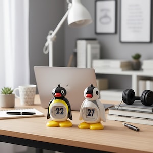 May include: Two penguin-shaped desk calendars. One is black and yellow, displaying '22', the other is white and grey, displaying '27'. They sit on a wooden desk with a laptop, headphones, and a pen. The desk is in a home office.