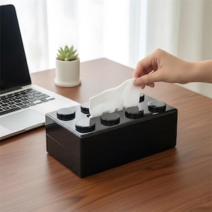 May include: A black tissue box, styled to look like a building block. The box has a rectangular shape with circular studs on top. A hand is pulling a white tissue from the opening. The box is on a wooden desk.