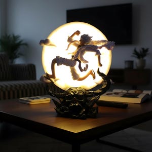 May include: A decorative lamp featuring a character leaping in front of a glowing, spherical light. The figure is in white clothing, with a dark base. The lamp is on a dark wooden table.