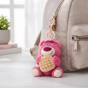 May include: A pink plush bear keychain with a waffle-shaped design. The bear has a smiling face, a dark pink nose, and a light pink body. It is attached to a rose gold keychain and a light gray backpack.