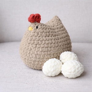 May include: A crocheted chicken in a light brown color with a red comb and yellow beak. Three white crocheted eggs are next to the chicken. The chicken is a decorative item.