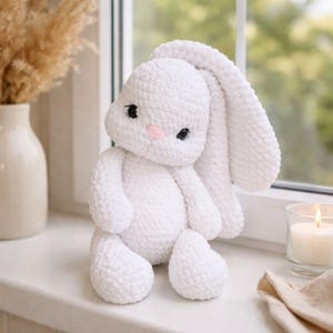 May include: A white crochet bunny toy with long ears, black eyes, and a pink nose. The soft toy is sitting on a white surface. The bunny is a handmade, cuddly toy.