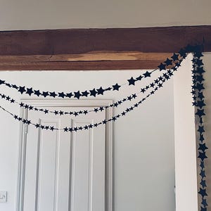 May include: Three strings of black star-shaped garland are draped across a doorway. The garland is strung across a wooden beam and hangs in front of a white door. The stars are evenly spaced, creating a festive, decorative look.