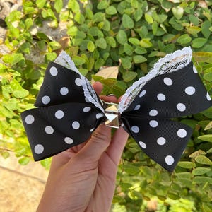 May include: A black hair bow featuring a white polka dot pattern and white lace trim along the edges. The center of the bow is adorned with a metallic silver pyramid stud, creating a structured look for this hair accessory piece.