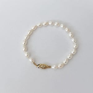 May include: A delicate pearl bracelet featuring oval-shaped pearls strung together. The bracelet is secured with a gold-colored clasp. The pearls are a creamy white color, and the bracelet is displayed on a white background.