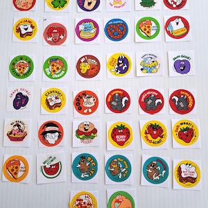 Puede incluir: Una colección de pegatinas vintage con ilustraciones y texto coloridos. Las pegatinas presentan varios personajes de dibujos animados, alimentos y frases como "Lucky Me!" y "Berry Good!" Las pegatinas están sobre un fondo blanco.