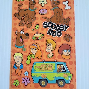 May include: A sheet of Scooby-Doo stickers featuring various characters and elements from the cartoon series. The stickers include images of Scooby-Doo, Shaggy, Fred, Daphne, Velma, and the Mystery Machine van, set against an orange background.