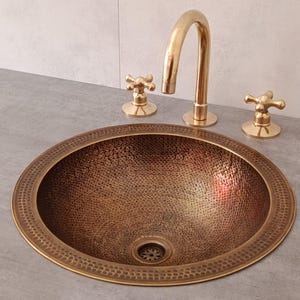 Può includere: Un lavabo ovale color rame martellato con bordo decorativo. È abbinato a un rubinetto color oro e rubinetti con maniglia a croce coordinati. I rubinetti sono montati su un piano di lavoro grigio.