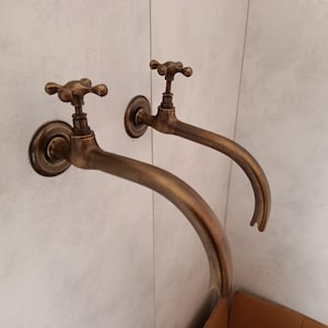 Op de afbeelding: Twee kranen in antieke bronzen stijl gemonteerd op een lichtgekleurde muur. Elke kraan heeft een kruisvormige handgreep en een gebogen tuit. De armaturen zijn gemaakt van bronskleurig metaal.