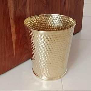 Puede incluir: Un cesto de basura de metal dorado con una textura martillada. El recipiente cilíndrico tiene una parte superior ligeramente ensanchada y una base redondeada. El cesto de basura está sobre un suelo de baldosas blancas, con un mueble de madera en el fondo.
