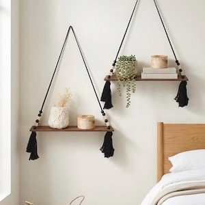 May include: Two hanging wooden shelves with black rope supports and tassels. Each shelf displays decorative items such as plants, candles, and a vase. The shelves are suspended from the ceiling, offering a stylish storage solution.
