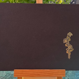 May include: A rectangular black card featuring a metallic gold foil illustration of a floral stem with multiple blossoms positioned on the right side. The card has a textured surface and a deckled edge on the right side, presenting a clean design.