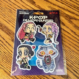 Peut inclure: Un ensemble de quatre écussons brodés représentant des personnages de K-Pop Demon Hunters et une créature sur une carte de support Netflix. Les écussons incluent trois personnages de style anime et un monstre bleu aux grands yeux jaunes.