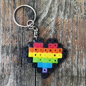 May include: A heart-shaped keychain constructed from fused plastic beads. The heart features horizontal stripes of red, orange, yellow, green, blue, and purple, all outlined by a border of black beads. A silver metal split ring is attached to the top.