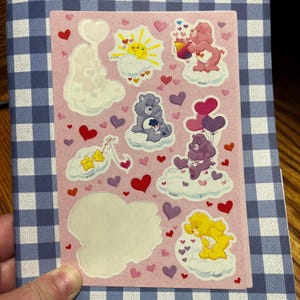 Puede incluir: Una hoja de pegatinas rectangular con fondo rosa claro que muestra varios personajes de Care Bears sobre nubes, incluyendo un oso rosa con un cubo, un oso morado con globos, un oso amarillo y un sol, rodeados por muchos corazones pequeños rojos y morados.