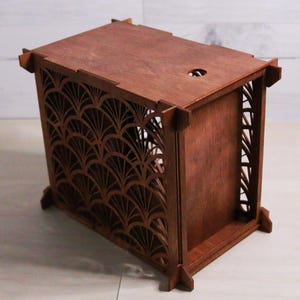 May include: A wooden box with a dark brown finish. The box features intricate, fan-shaped cutouts on one side and decorative corner accents. The top has a small circular opening. The box is likely for storage or display.