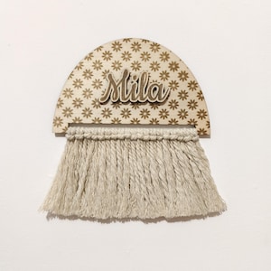Puede incluir: Adorno de pared de madera con el nombre "Mila" en escritura cursiva. La parte superior es un semicírculo con un estampado floral, y la parte inferior presenta una franja de macramé beige.
