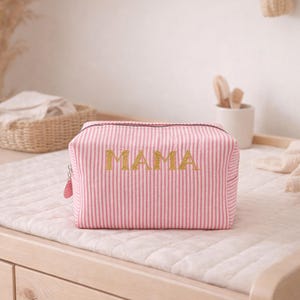 Op de afbeelding: Roze en wit gestreepte make-uptas met het woord "MAMA" in gouden glitterletters. De tas heeft een ritssluiting en een roze ritstrekker. De tas ligt op een wit gewatteerd aankleedkussen.