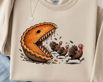 May include: A sand-colored crewneck sweatshirt featuring a graphic of a large, toothy pumpkin pie chasing three turkeys. The pie has a golden-brown crust and sharp teeth. The sweatshirt has a Gildan label at the inner neckline and long sleeves.