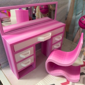 May include: A miniature pink and white vanity desk with a rectangular mirror and three drawers on each side, accompanied by a matching pink S-shaped chair. The vanity features white drawer fronts and white handles, with a central open storage space.