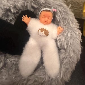 May include: A doll dressed in a white, fluffy onesie with a hedgehog applique, lying in a gray, plush bed. The doll has a white headband with a pink bow. The doll's arms are outstretched.