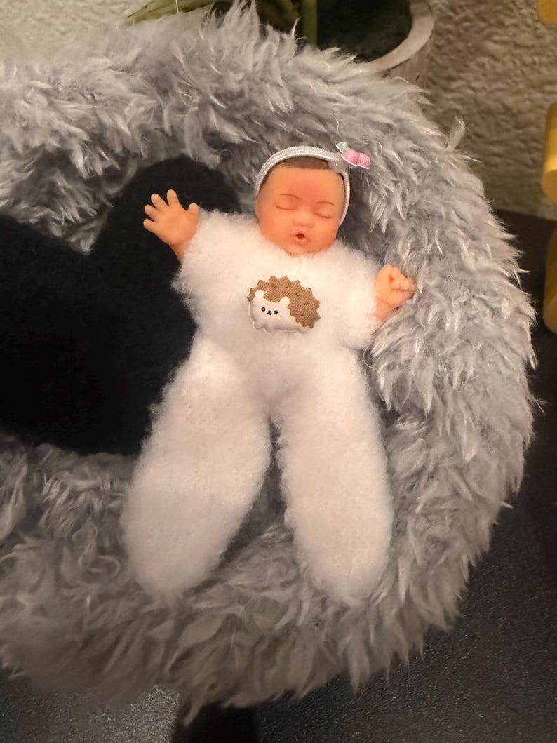 May include: A doll dressed in a white, fluffy onesie with a hedgehog applique, lying in a gray, plush bed. The doll has a white headband with a pink bow. The doll's arms are outstretched.