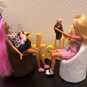 May include: Two Barbie dolls are seated, one with pink and blue hair holding a baby doll, the other with blonde hair holding a brown doll. The dolls are in a room with a white wall and a black surface. The dolls are wearing casual clothes.