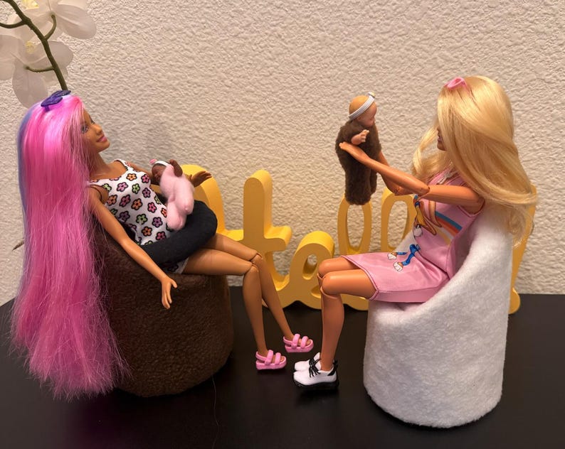 May include: Two Barbie dolls are seated, one with pink and blue hair holding a baby doll, the other with blonde hair holding a brown doll. The dolls are in a room with a white wall and a black surface. The dolls are wearing casual clothes.