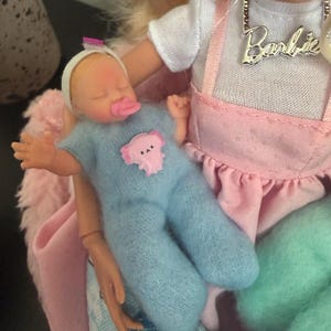 May include: A baby doll wearing a blue onesie with a pink bear applique and a white headband. The doll is held by a Barbie doll wearing a white shirt and pink overalls. A silver necklace with the name "Barbie" is visible.