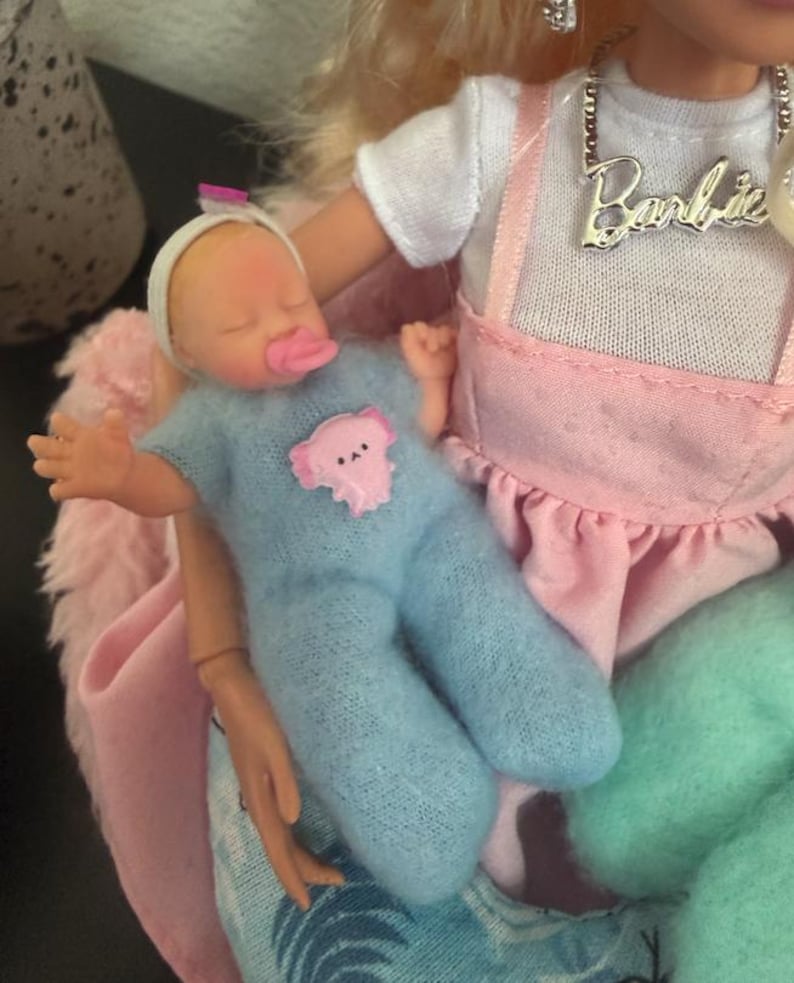 May include: A baby doll wearing a blue onesie with a pink bear applique and a white headband. The doll is held by a Barbie doll wearing a white shirt and pink overalls. A silver necklace with the name "Barbie" is visible.