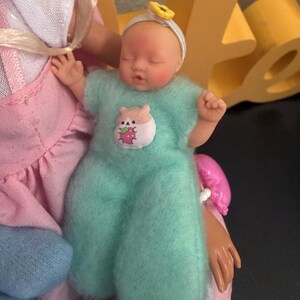 May include: A small baby doll wearing a light blue fuzzy onesie with a hamster and strawberry patch. The doll has a white headband with a yellow flower and is held by a larger doll in a pink dress.