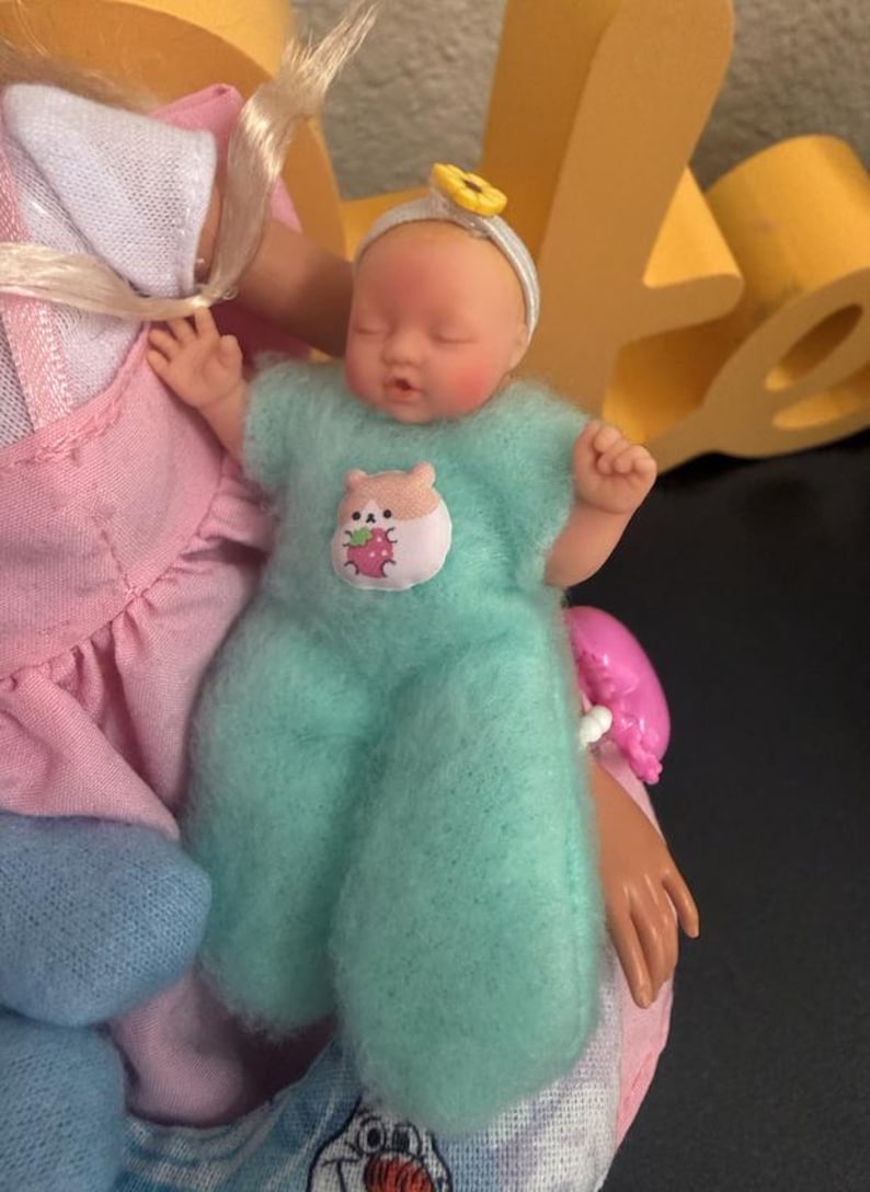 May include: A small baby doll wearing a light blue fuzzy onesie with a hamster and strawberry patch. The doll has a white headband with a yellow flower and is held by a larger doll in a pink dress.