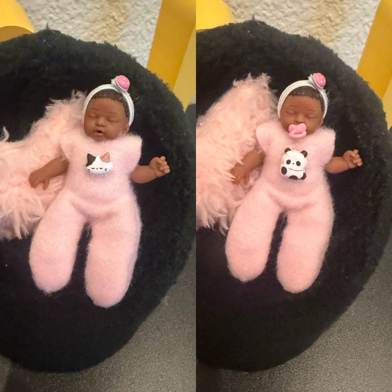 May include: A small doll wearing a pink onesie with a cat graphic, a white headband, and a pink pacifier. The doll is lying on a black surface with a pink fluffy blanket. The doll's skin is brown.