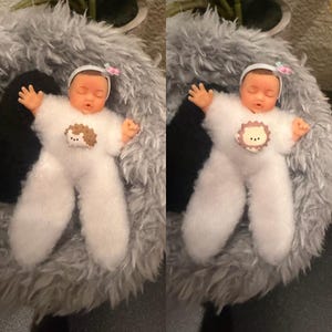 May include: A doll dressed in a white, fluffy outfit with a hedgehog applique, lying on a gray, furry surface. The doll has a white headband with a pink bow and is posed with arms outstretched. The doll's skin is a light peach color.